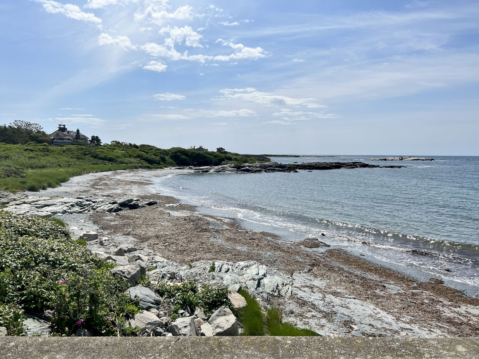 A scenic rocky coastline with waves, green shrubs, and a lighthouse-like house under a bright, cloud-filled blue sky.