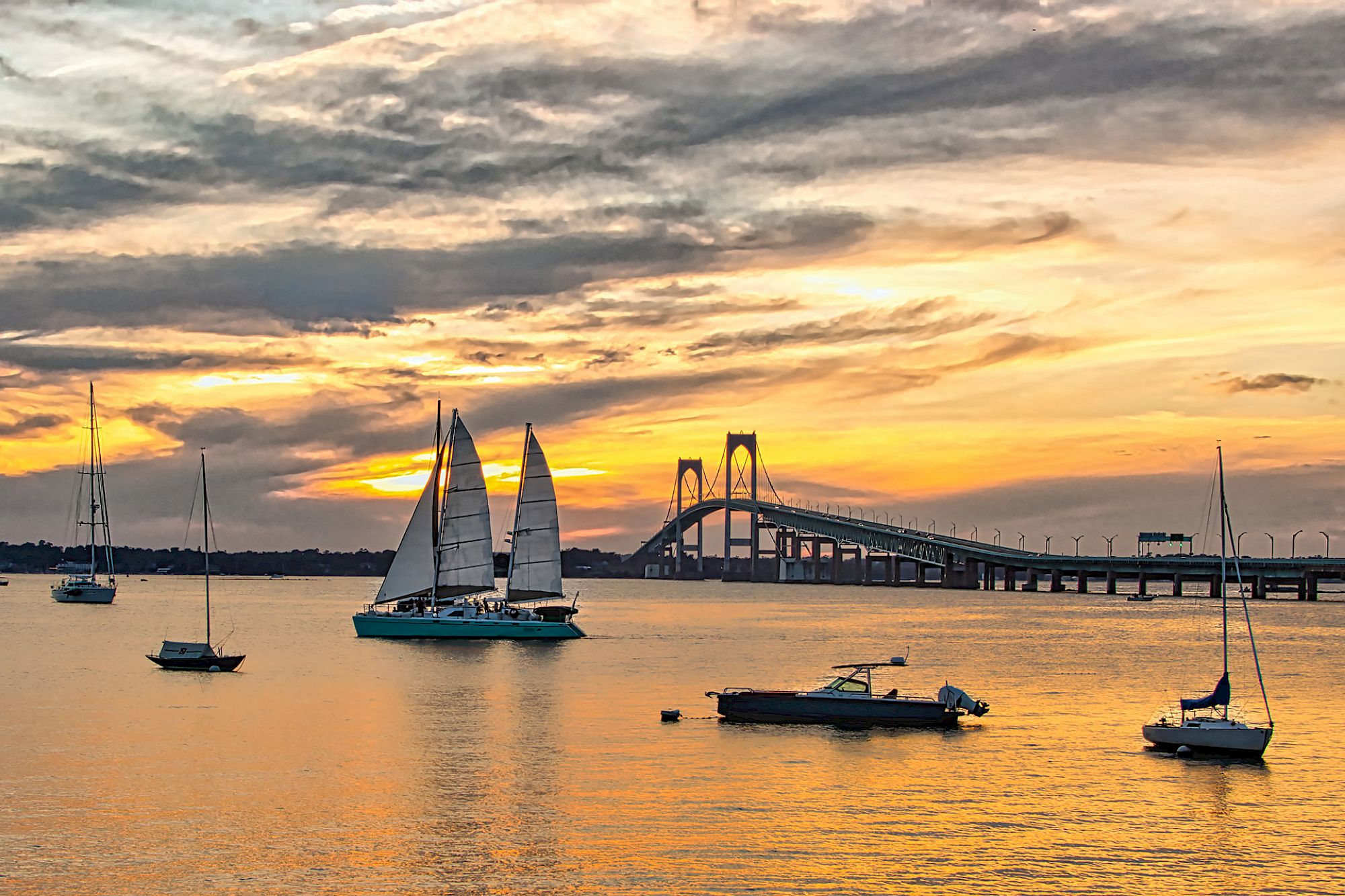 A scenic sunset view of a bay with sailboats and a bridge in the background against a dramatic sky.