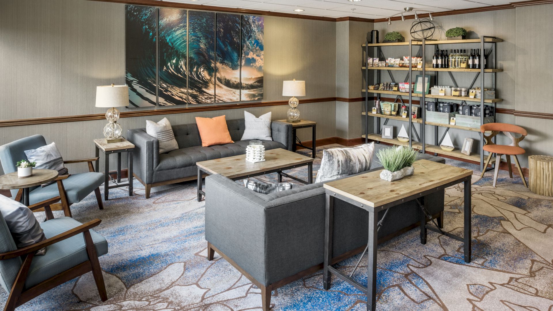 A cozy lounge with gray sofas, a wave-themed wall art, bookshelves, and plants on tables and shelves, with a patterned carpet.