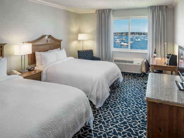 The image shows a hotel room with two beds, a TV, a desk, and a window with a view of boats on the water.