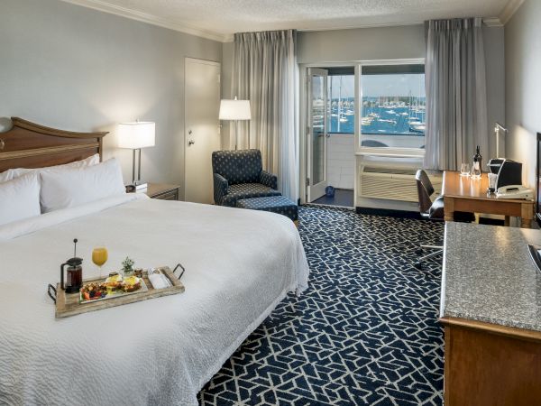 A hotel room with a large bed, desk, TV, armchair, and a tray of breakfast on the bed. Balcony shows a marina view.