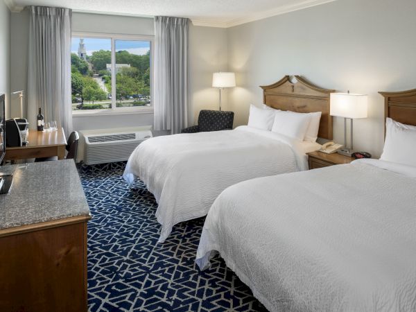 The image shows a hotel room with two beds, a window view, a TV, desk, and lamps on nightstands, creating a cozy and clean atmosphere.
