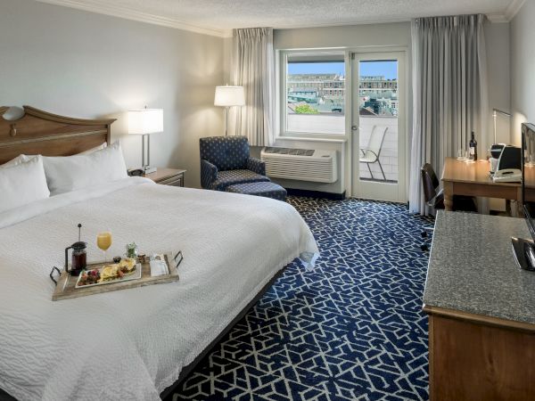 A hotel room with a large bed, TV, desk, armchair, and a tray with drinks. There's a window with a view. Navy patterned carpet adds style.