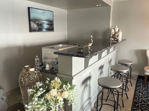 The image shows a home bar area with stools, decorative vases, and flowers, set against a gray wall with a picture hanging above.