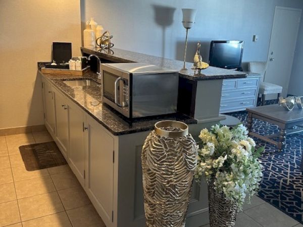 A kitchen area with a countertop, microwave, large decorative vases, flowers, and a living room with a TV in the background.