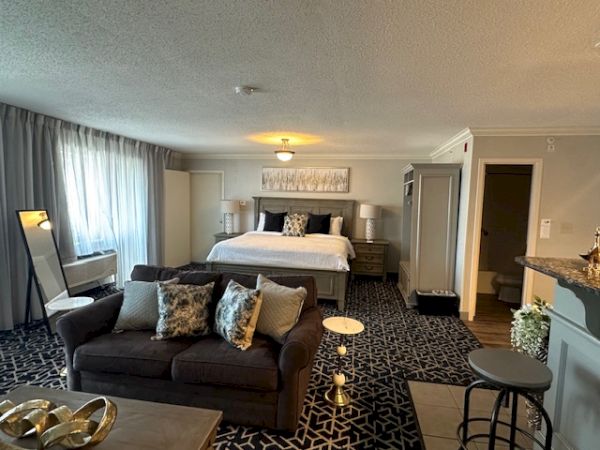 A modern hotel room with a bed, sofa, chair, and bar stools. There’s a patterned carpet and soft lighting.