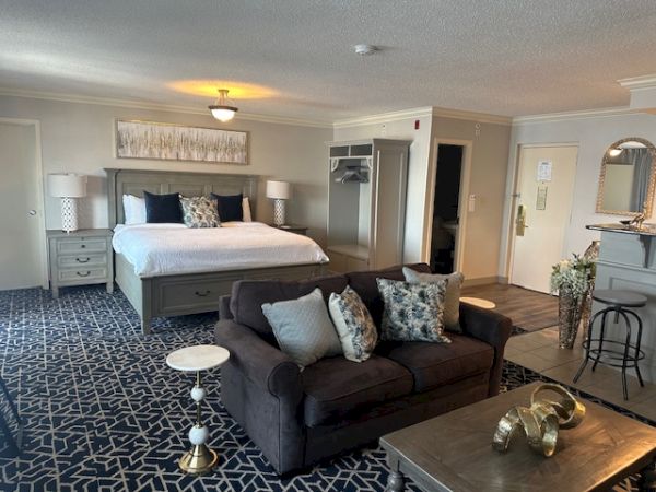 A spacious hotel room with a bed, sofa, and coffee table. It features modern decor, a patterned rug, and neutral color tones.