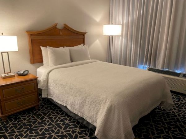 A neatly made bed with white linens is in a hotel room, featuring wooden furniture, lamps, and patterned carpet flooring.