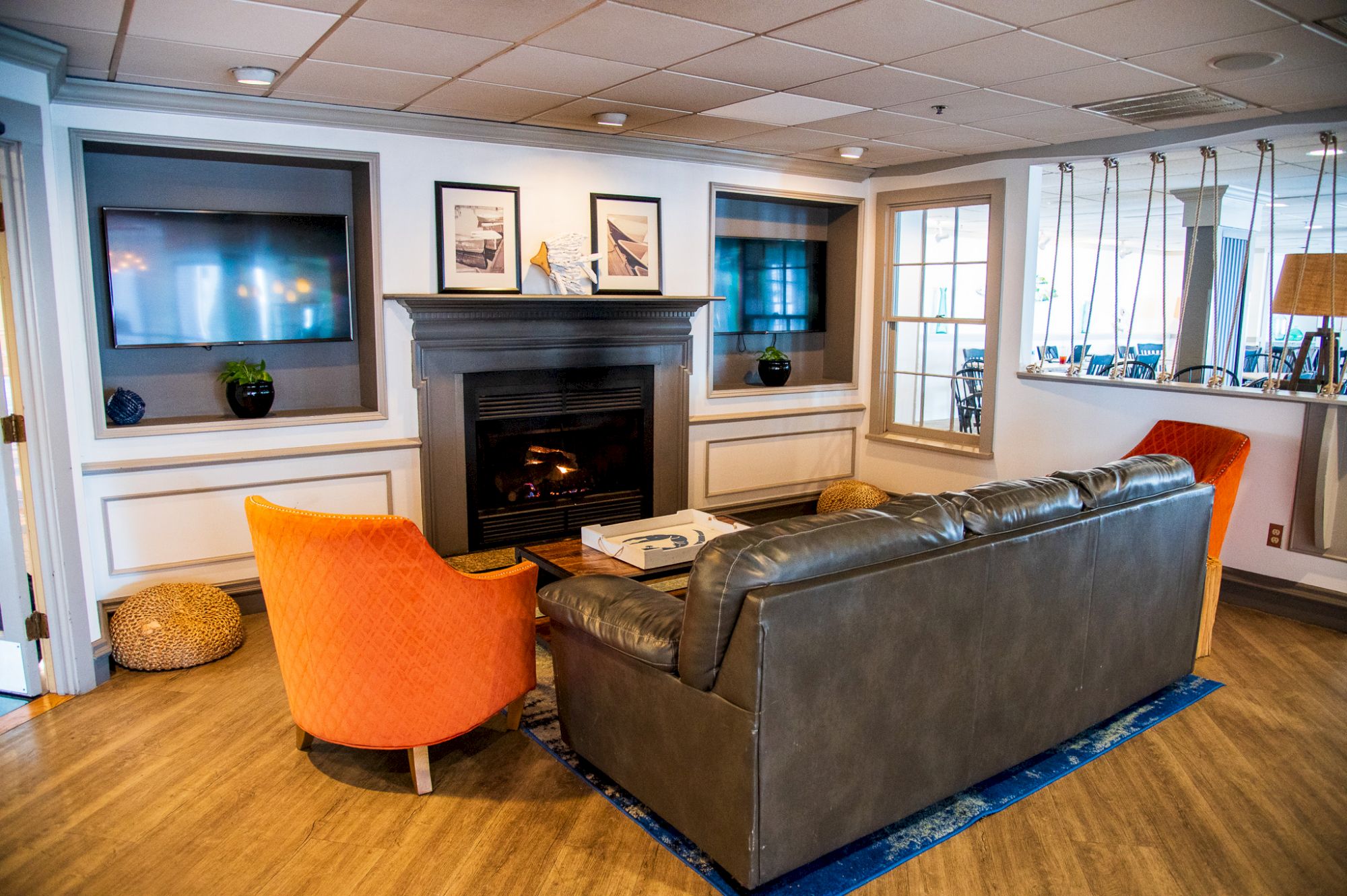A cozy living room with a fireplace, two TVs, a gray couch, orange chairs, and framed art. The floors are wood, adding warmth.