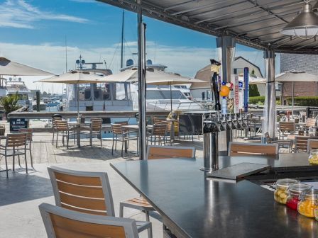 An outdoor seating area by a marina with boats, tables, chairs, and a bar setup under umbrellas on a sunny day.