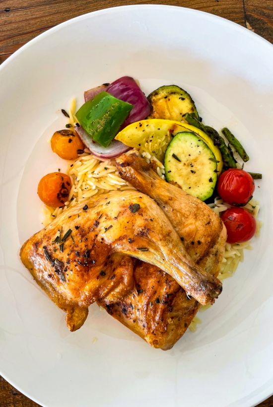 Grilled chicken served with roasted vegetables including zucchini, bell peppers, cherry tomatoes, and onions, over a pasta bed.