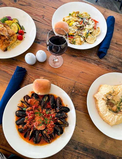 The image shows a table with seafood dishes, pasta, bread, wine, and napkins on a wooden surface.