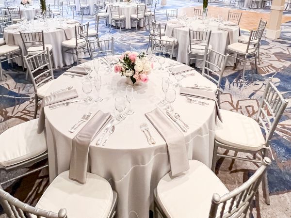 The image shows an elegant banquet hall set up with round tables, white tablecloths, silver chairs, and flower centerpieces.