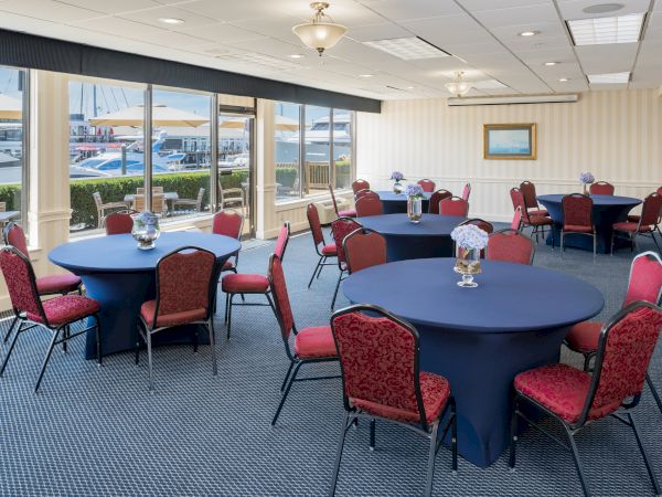 A conference room with round tables covered in blue tablecloths and red chairs, overlooking a view of boats outside large windows.