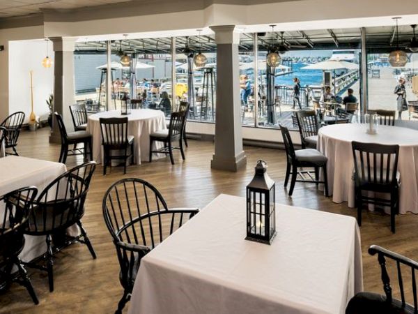 A restaurant with white tablecloths and black chairs, featuring large windows overlooking a waterfront.
