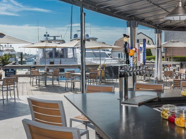 An outdoor restaurant bar with wooden chairs overlooks a marina with docked boats. The area is shaded by umbrellas and a metal awning.
