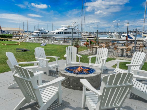 A marina scene with white chairs around a fire pit, overlooking boats and yachts on a sunny day. There are tables and a hammock nearby.