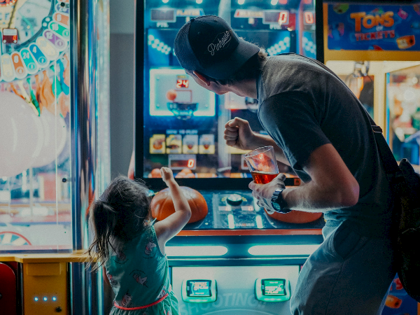 A man helps a young girl at an arcade claw game; she reaches toward the prize while he guides her, both focused on the screen.
