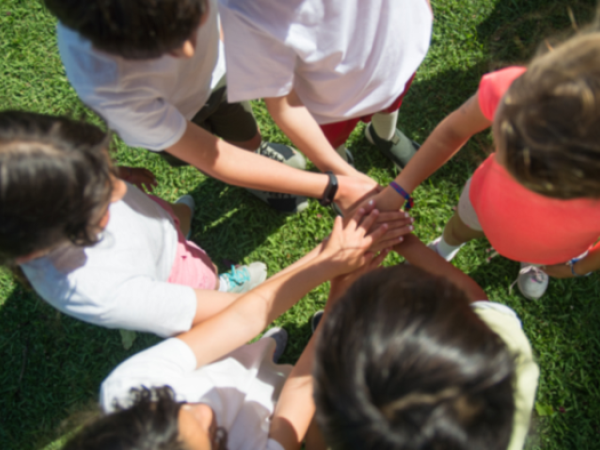 A group of children standing in a circle, each placing their hand in the center on top of the others, on a grassy field.