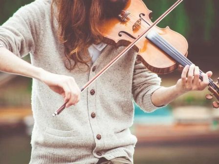 A person playing a violin outdoors, wearing a sweater, and focusing on the instrument.