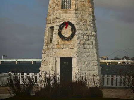 A stone lighthouse is decorated with a holiday wreath, standing near the water with a cloudy sky in the background.