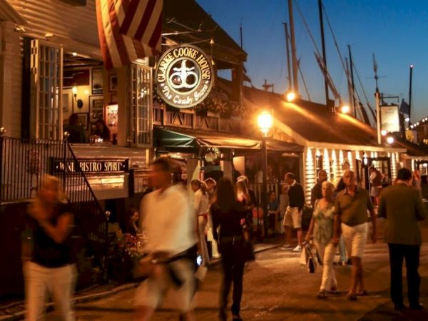 A lively street scene at night with people walking near illuminated shops and a restaurant. Sailboats are visible in the background under a clear sky.