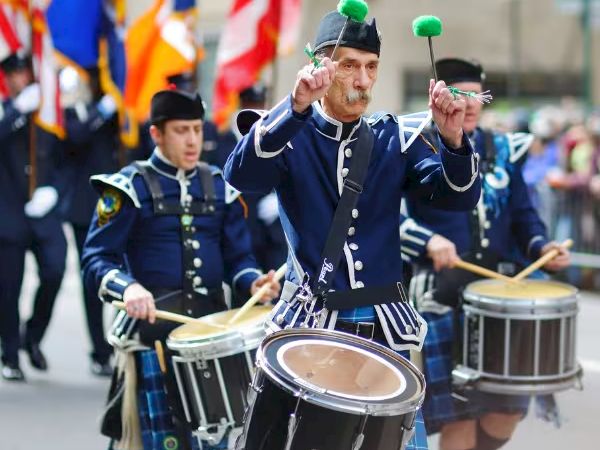 A marching band in traditional uniforms playing drums during a parade, with flags waving in the background.