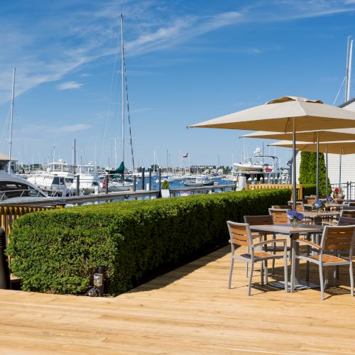 Outdoor dining area with tables, umbrellas, and chairs on a wooden deck overlooking a marina with boats under a clear blue sky.