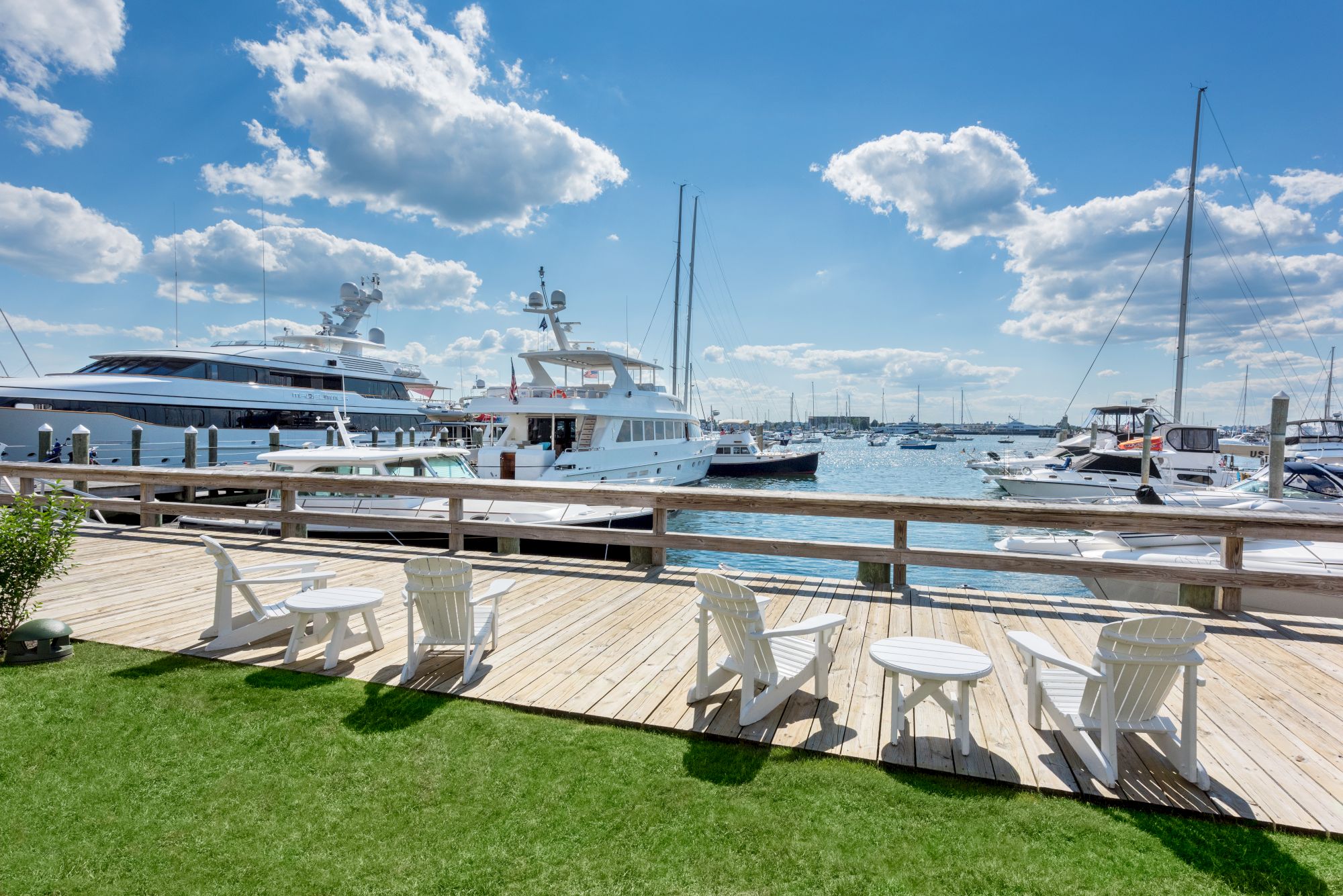 The image shows a marina with several yachts docked. White chairs and tables are set on a wooden deck overlooking the water under a blue sky.