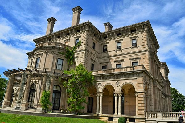 A grand, historic mansion with detailed architecture, arched windows, and lush greenery under a bright blue sky.
