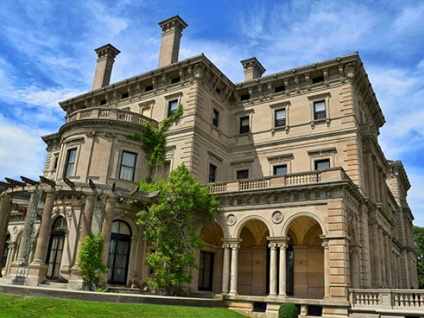 A grand, historic mansion with detailed architecture, arched windows, and lush greenery under a bright blue sky.