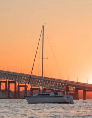 A suspension bridge silhouetted against a sunset sky with a sailboat anchored on the water in the foreground.