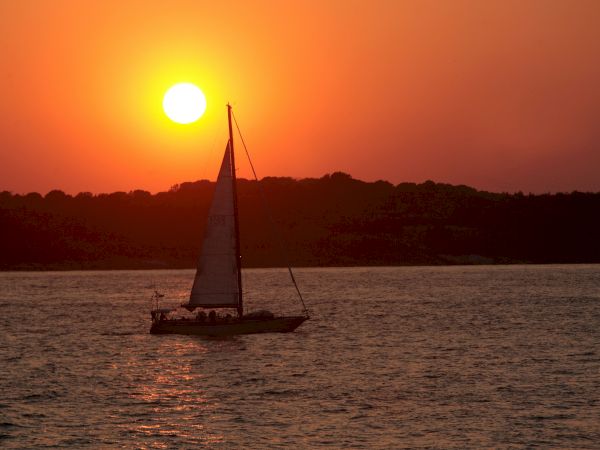 A sailboat is silhouetted against a vibrant orange sunset over a calm body of water, with hills in the background.
