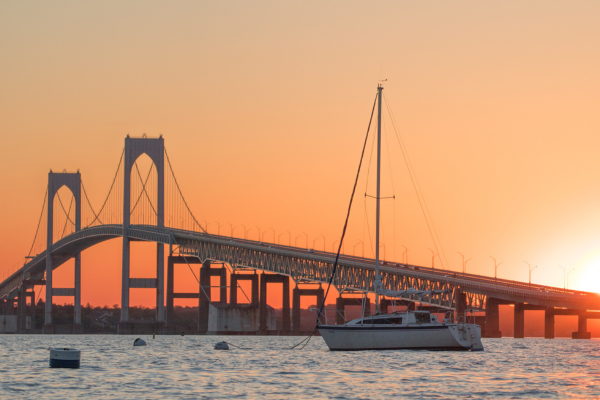 A sailboat on the water with a bridge in the background is silhouetted against an orange sunset sky.