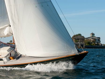 A sailboat glides on the water with white sails billowing, near a shoreline with buildings and a flag in the background.