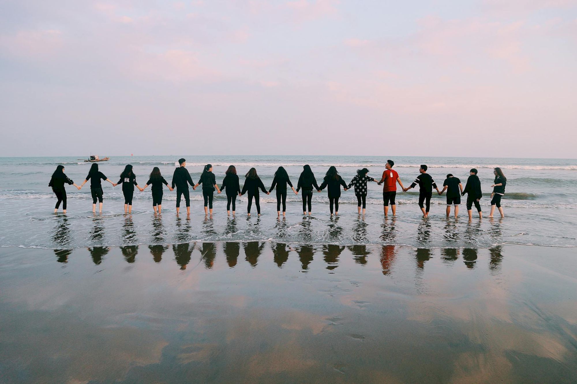 A group of people holding hands stands in a line on the beach, reflected in wet sand, facing the sea at sunset.