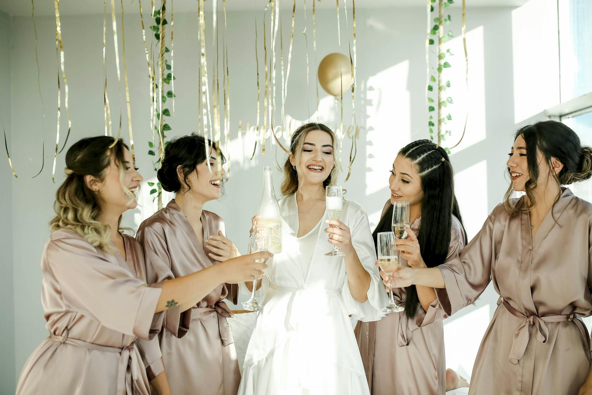 A group of women in matching robes and the bride are joyfully toasting with champagne under hanging decorations in a bright room.