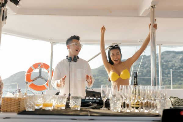 A person DJing on a boat with a woman in a yellow bikini raising her arms, surrounded by glasses and a scenic backdrop.
