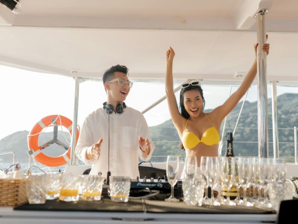 A person DJing on a boat with a woman in a yellow bikini raising her arms, surrounded by glasses and a scenic backdrop.