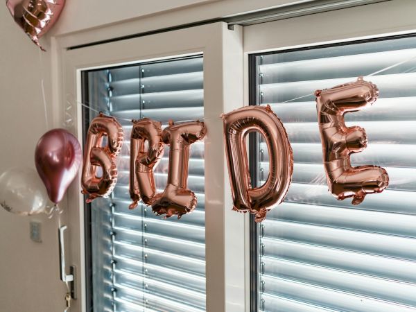 The image shows metallic balloon letters spelling "BRIDE" with additional balloons, in front of a window with blinds.