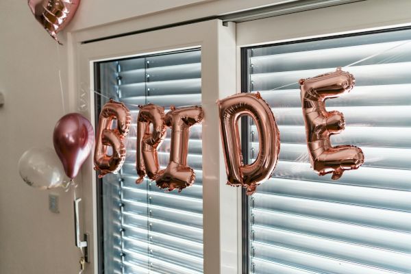 The image shows metallic balloon letters spelling "BRIDE" with additional balloons, in front of a window with blinds.
