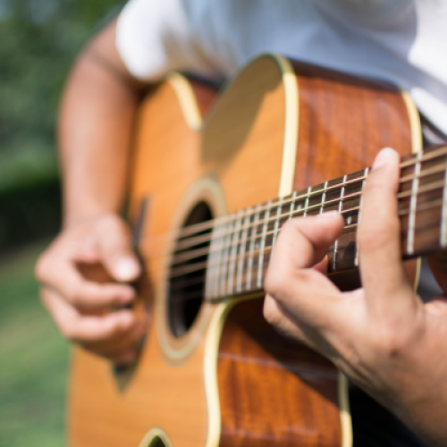 A person is playing an acoustic guitar outdoors, with a focus on their hands and the guitar's strings and body.