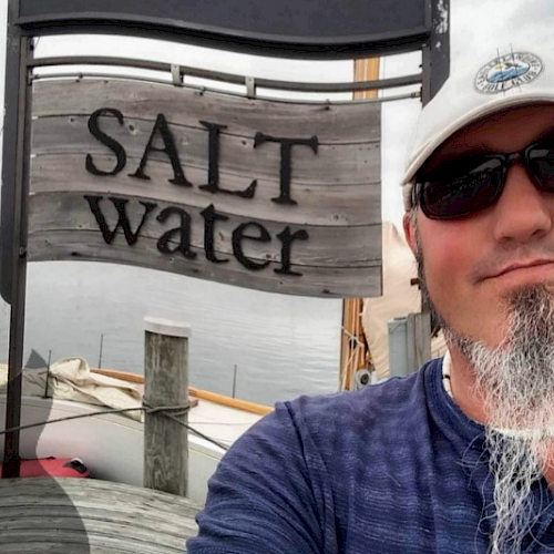 A person taking a selfie in front of a wooden sign reading "SALT water" near a waterfront, wearing sunglasses and a cap.