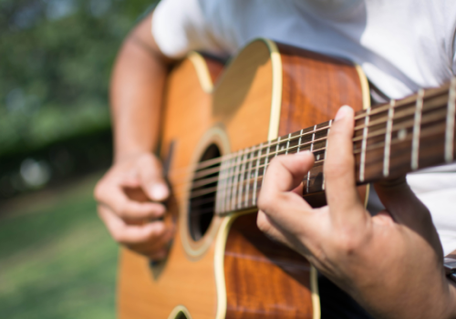 A person is playing an acoustic guitar outdoors. The focus is on the guitar and the hands, with blurred greenery in the background.