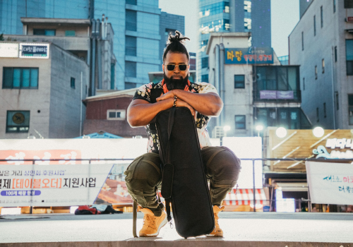 A person in sunglasses and casual attire kneels with a guitar case in an urban setting, with buildings and signs in the background.