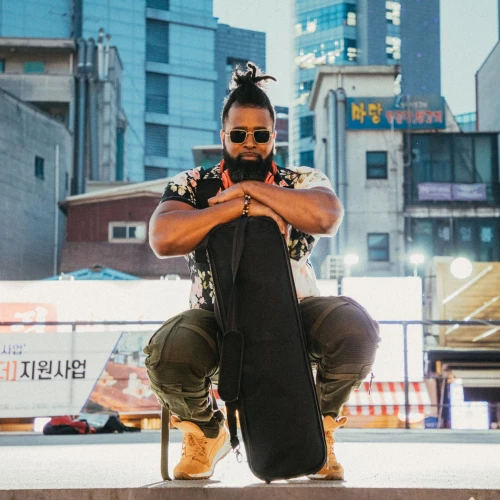 A person in sunglasses and casual attire kneels with a guitar case in an urban setting, with buildings and signs in the background.