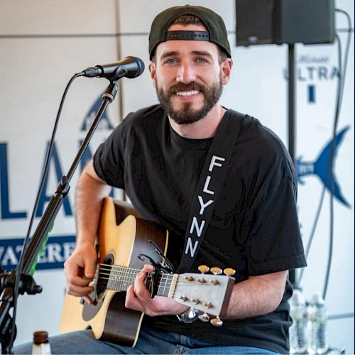 A smiling man in a cap plays an acoustic guitar onstage, singing into a mic during a live performance.