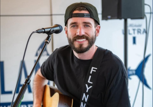A man is playing an acoustic guitar and singing into a microphone. He is wearing a cap and a "FLYNN" shirt, seated indoors.