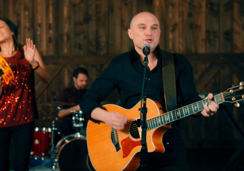 A band performing, featuring a guitarist singing at a mic, a woman with a tambourine, and a drummer in the background.
