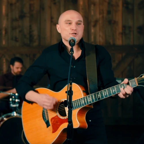 A band performing, featuring a guitarist singing at a mic, a woman with a tambourine, and a drummer in the background.